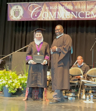 A picture of me during my high school graduation, I'm on the stage having just received my certificate and standing next to the principle at the time. I'm wearing a black graduation gown with purple accents.
